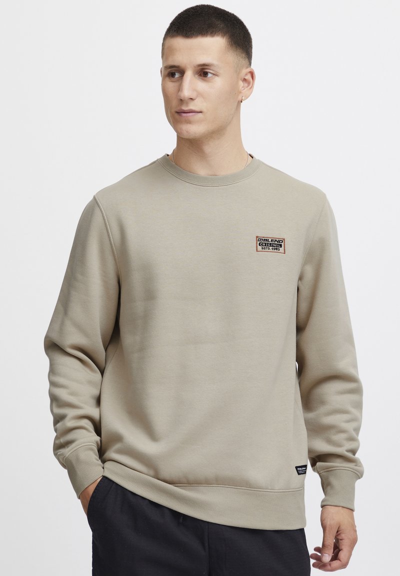 Sudadera beige de algodón suave, con cuello redondo, puños acanalados y una pequeña etiqueta en el pecho. Acabado de tela texturizada.