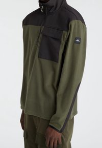 Fleece trui in olijfgroen met een zwart bovenpaneel, halve ritssluiting bij de nek, borstzak en logovlaggetje op de mouw. Soepele textuur, getailleerde pasvorm.