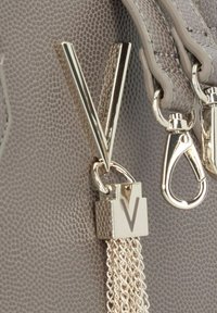 Detalj av en grå texturerad läderväska med en guldfärgad "V"-berlock och lås, med silverkedjeackenter och metalliska beslag.