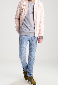 Light pink bomber jacket, gray graphic sweatshirt, distressed light blue jeans with scribble patterns, and brown sneakers with yellow accents.