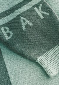 Audinio mėlynos ir tamsiai žalios spalvų pledukas, ant kurio užrašyti „BAK“ šviesesne spalva. Jame yra raštuotas kraštas ir tekstūruotas paviršius.