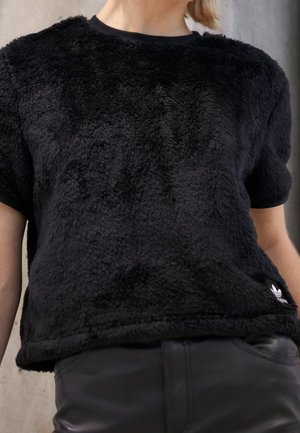 Personne portant un pull noir à manches courtes en tissu duveteux et un pantalon en cuir noir, montrant le logo Adidas près de l'ourlet.