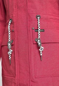 Nærbillede af den røde jakke foran med to lappe lommer, hvide og sorte snore med metalspidser og metaltrykknapper.