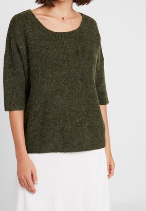 Woman wearing a fuzzy dark green knit sweater with three-quarter sleeves and a white skirt, standing against a plain light background.