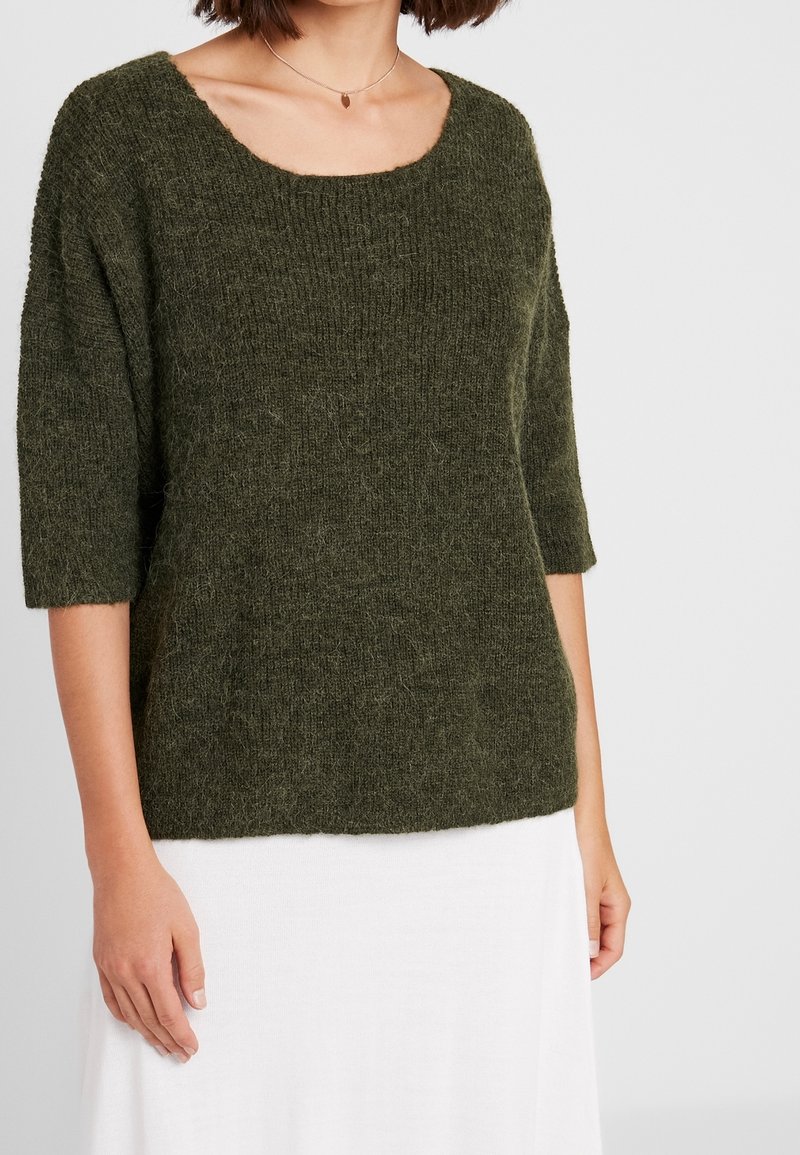 Femme portant un pull en maille vert foncé duveteux à manches trois-quarts et une jupe blanche, debout devant un fond clair uni.