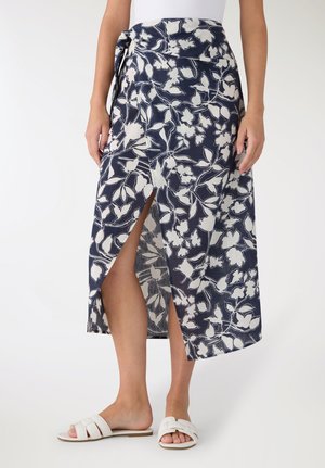 Donna che indossa una gonna a portafoglio blu navy con stampa floreale bianca e spacco frontale, abbinata a sandali bianchi a ciabatta.