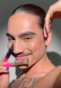 Två NYX kosmetikprodukter: en rosa och svart mascara, samt en brun foundationtub som hålls framför en person med en gul eyeliner-accent.
