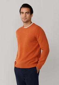 Pomarańczowy sweter dzianinowy z okrągłym dekoltem oraz ściągaczami przy mankietach i hemie. Noszony na białą koszulę, w połączeniu z ciemnogranatowymi spodniami.