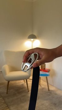 Srebrna zaponka pasu, držana v roki, s črnim trakom, ki se spušča navzdol. V ozadju je svetel stol in svetilka.