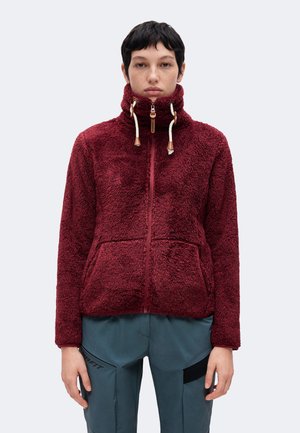 Persoon met kort donker haar, die een bordeauxrode fleecejas met rits en blauwe broek draagt, voor zich uitkijkend tegen een effen lichte achtergrond.