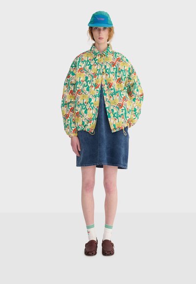 Femme portant une veste colorée à motifs floraux, une robe en jean bleu, une casquette turquoise, des chaussettes blanches à rayures vertes et des chaussures marron, debout devant un fond blanc.