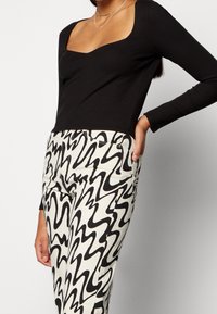 Musta ribbitty pitkähihainen toppi, jossa on neliönmuotoinen kaula-aukko, yhdistettynä valkoisiin housuihin, joissa on voimakkaat mustat aaltoilevat kuviot. Sujuva kangas, istuva muotoilu.