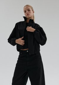 Kobieta z blond włosami, ubrana w czarną krótką kurtkę nad czarną bluzką oraz czarnymi szerokimi spodniami, na jednolitym szarym tle.