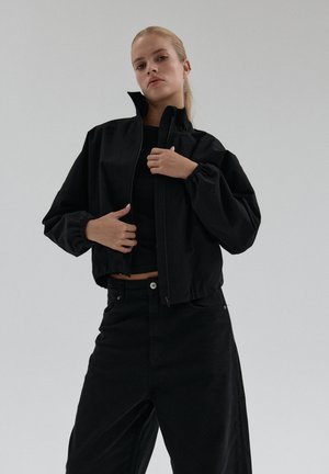 Kobieta z blond włosami, ubrana w czarną krótką kurtkę nad czarną bluzką oraz czarnymi szerokimi spodniami, na jednolitym szarym tle.