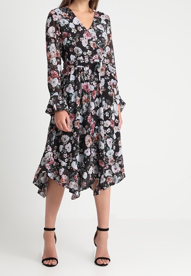 Robe fleurie avec des manches longues et superposées et un décolleté en V. Le tissu noir présente des fleurs roses, blanches et vertes. Ourlet en forme de mouchoir et taille ceinturée.
