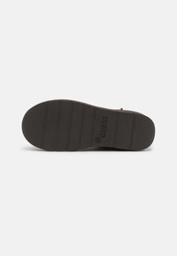 Suela de zapatilla marrón oscuro con surcos horizontales para tracción. Presenta el logotipo "GUESS" en relieve y detalles de tamaño. Material de goma.