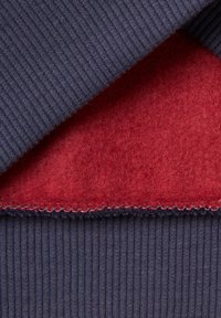 Tela de punto acanalado azul navy superpuesta con un suave fieltro rojo. Detalle de costura fina visible a lo largo del borde donde se encuentran los dos materiales.