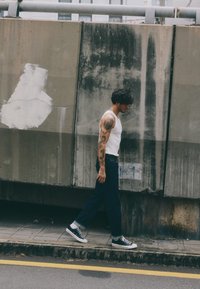 Canottiera bianca e jeans blu scuro, sneakers bianco e nere, con tatuaggi visibili sul braccio, che cammina accanto a un muro di cemento.