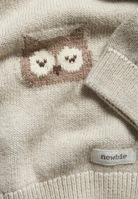 Beige neulottu neulepaita, jossa on ruskea pöllökuvio etupuolella ja ala-reunaan ommeltu "newbie"-tunnus.