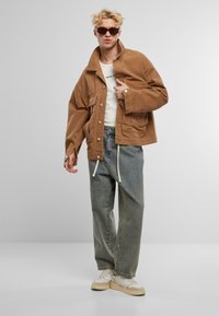 Braune Cordjacke mit spitzem Kragen und Fronttaschen, kombiniert mit hellblauen, baggy Jeans und kremfarbenen Sneakern.