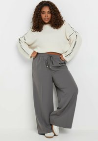 Krämfärgad stickad tröja med svarta sidostygn, parat med grå vida byxor och vita sneakers. Avslappnad passform, mjuk textur, avslappnad stil.