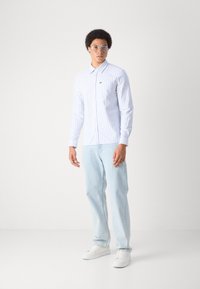 Weißes Langarmhemd mit hellblauen vertikalen Streifen, Kragen und Knöpfen, kombiniert mit hellblauen Jeans und weißen Sneakers.