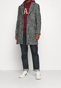 Soprabito grigio a spina di pesce con una vestibilità sartoriale, indossato sopra una felpa con cappuccio color bordeaux. Abbinato a jeans scuri e sneakers bianche. Tessuto liscio, maniche lunghe.