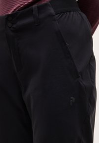 Κοντινό πλάνο μαύρου παντελονιού με πλαϊνή τσέπη με φερμουάρ και κλείσιμο με δύο κουμπιά, φορεμένο με μπορντό τοπ.