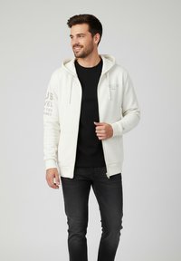 Cremefarbene Zip-Hoodie mit Kängurutasche, Kapuze mit Zugband und Textdetails am linken Ärmel, getragen über einem schwarzen Shirt und dunklen Jeans.