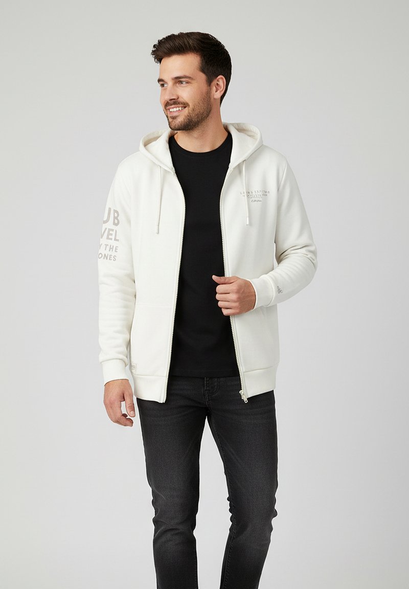 Cremefarbene Zip-Hoodie mit Kängurutasche, Kapuze mit Zugband und Textdetails am linken Ärmel, getragen über einem schwarzen Shirt und dunklen Jeans.