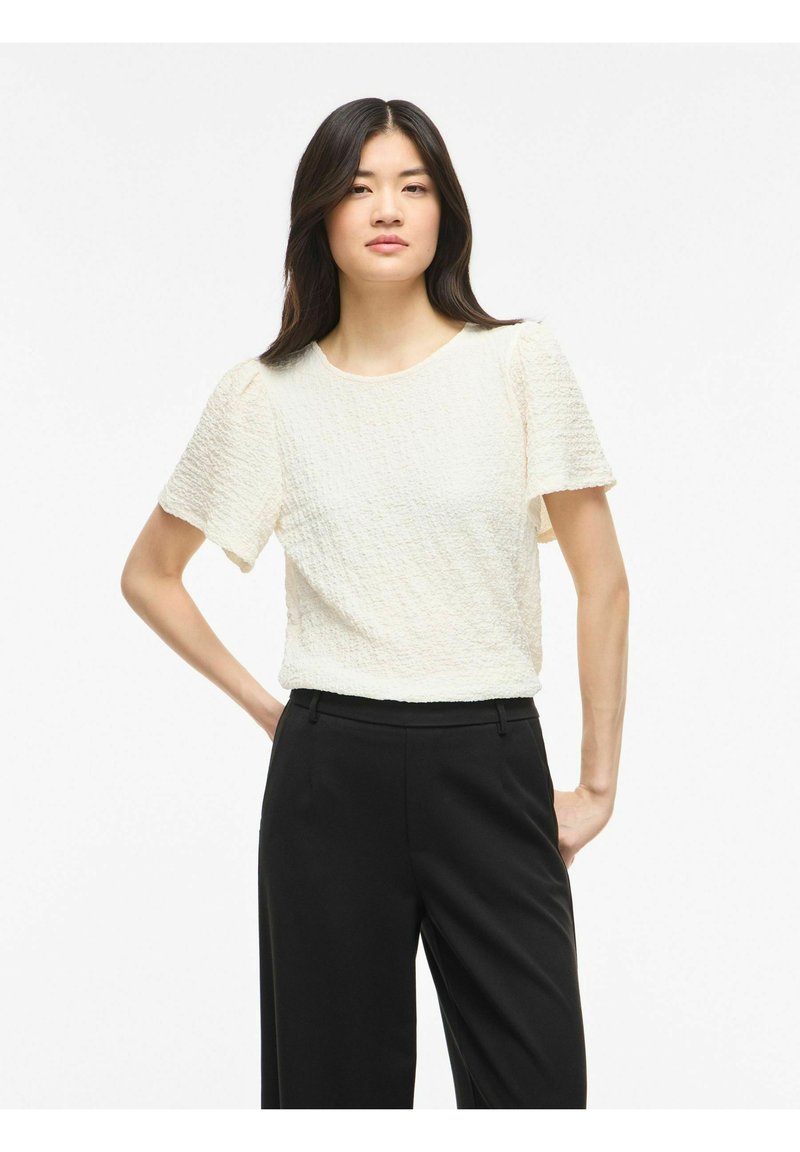 Femme aux longs cheveux foncés portant un haut blanc texturé à manches courtes et un pantalon noir, debout, les mains sur les hanches, sur fond blanc.