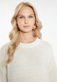 Maglione bianco lavorato a maglia con un ampio scollo. Il modello indossa orecchini rotondi con accenti dorati e rossi. Capelli acconciati in onde morbide.