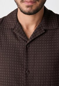 Camisa marrón abotonada con un patrón geométrico texturizado, cuello abierto y dos botones oscuros en la parte delantera.