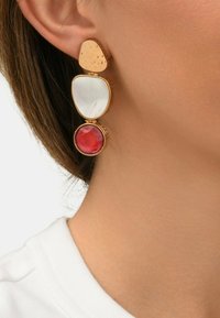 Boucles d'oreilles dorées présentant trois formes géométriques : un haut texturé en or, un milieu iridescent blanc et un élément circulaire rouge en bas.