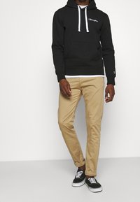 Black hoodie with white drawstrings, logo on the chest, paired with beige pants and black sneakers with white soles.