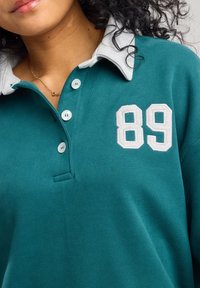 Maglione verde acqua con colletto grigio chiaro, dotato di tre bottoni e ricamo "89" in bianco. Texture morbida con un design sportivo.