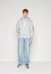 Felpa grigia con tasca frontale e logo, abbinata a jeans dritti blu chiaro. Le sneakers presentano accenti blu e bianchi.