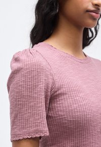 Ριμπ τοπ με κοντά μανίκια σε μωβ απόχρωση με κυματιστό ντεκολτέ. Η υφή του υφάσματος και οι φουσκωτοί ώμοι αναδεικνύουν το σχέδιο.