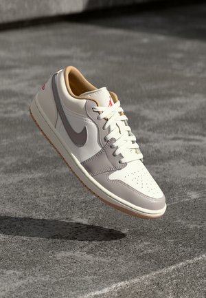 Białe i jasnoszare niskie buty sportowe z gumową podeszwą i czerwonym logo, pokazane w powietrzu nad teksturowaną, szarą powierzchnią.