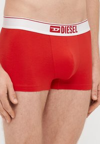 Sarkanas vīriešu bokseršorti no gludas auduma ar baltu elastīgu jostu, uz kuras redzams sarkans "DIESEL" logotips, un cieši pieguļošu dizainu.