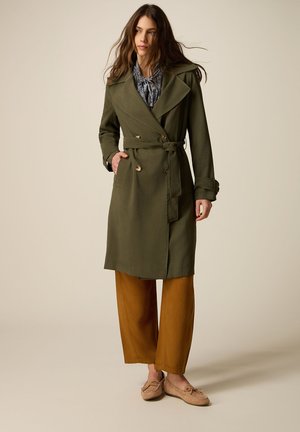 Donna che indossa un trench verde oliva con cintura, pantaloni marroni a gamba larga, mocassini beige e una camicia a fantasia, in piedi davanti a uno sfondo neutro.