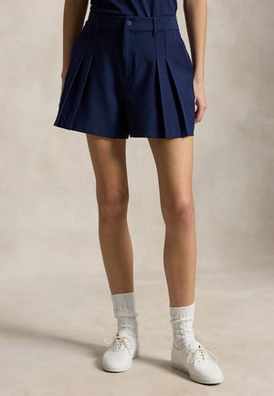 Kvinne iført plisserte marineblå shorts, hvite høye sokker og hvite snøresko, stående mot en nøytral bakgrunn.