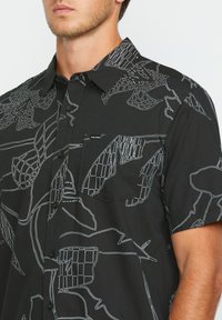 Camisa negra de manga corta con dibujos botánicos en líneas blancas, cuello abotonado, bolsillo en el pecho y etiqueta de logo cerca del bolsillo.