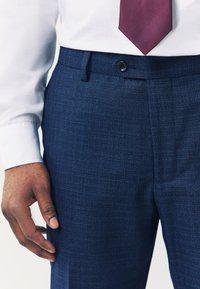 Dunkelblaue Anzughose aus leicht strukturiertem Stoff mit einem vorderen Knopfverschluss und Taschen, kombiniert mit einem weißen Hemd und einer bordeauxfarbenen Krawatte.
