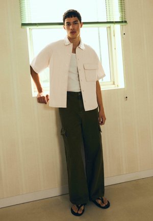 Fiatal férfi, aki egy ablak melletti falnak dőlve fehér rövid ujjú ingben, fehér trikóban, sötétzöld cargo nadrágban és papucsban áll.