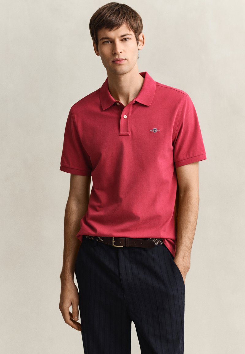 Jeune homme portant un polo rouge et un pantalon à fines rayures foncé avec une ceinture marron, debout devant un fond beige uni.