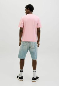 Helles rosa T-Shirt mit kurzen Ärmeln, lockere Passform, kombiniert mit hellblauen Denim-Shorts, schwarzen Sneakers und weißen Socken mit schwarzen Streifen.