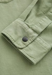 Light green textured fabric close-up featuring a folded sleeve with a black snap button detail and flat stitching.