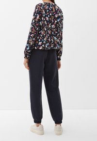 Blusa a fantasia floreale di un blu navy scuro con maniche lunghe e dettaglio a chiave nella parte posteriore; abbinata a pantaloni della tuta scuri e sneakers chiare.