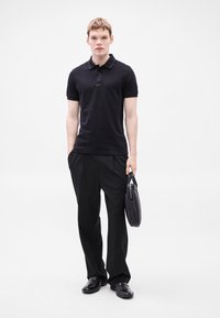 Melns polos krekls ar apkakli un logotipu, melnas bikses un spīdīgas melnas kurpes. Tur melnu portfeli. Vienkāršs balts fons.